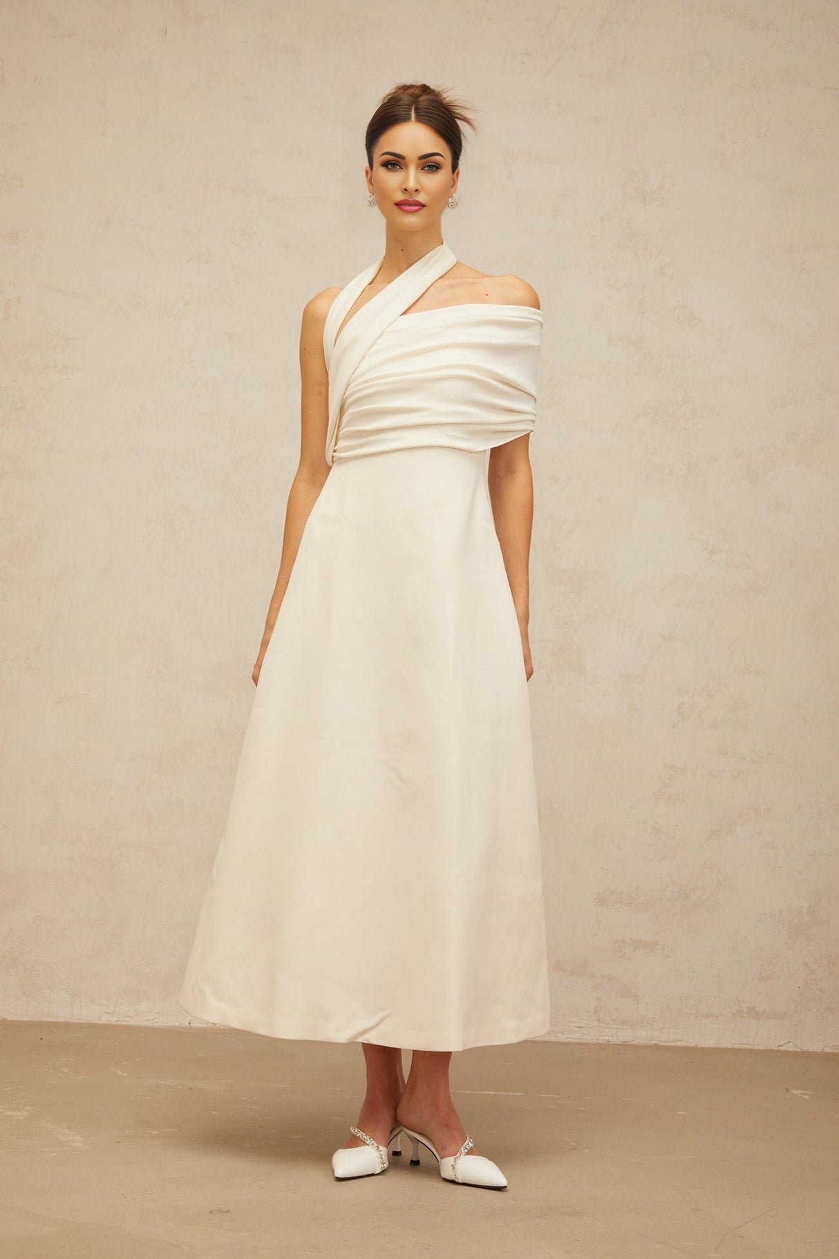 A woman wearing a long creamwhite dress with an asymmetric neckline and hemline standing against a plain background