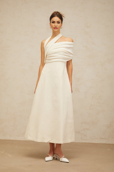 A woman wearing a long creamwhite dress with an asymmetric neckline and hemline standing against a plain background