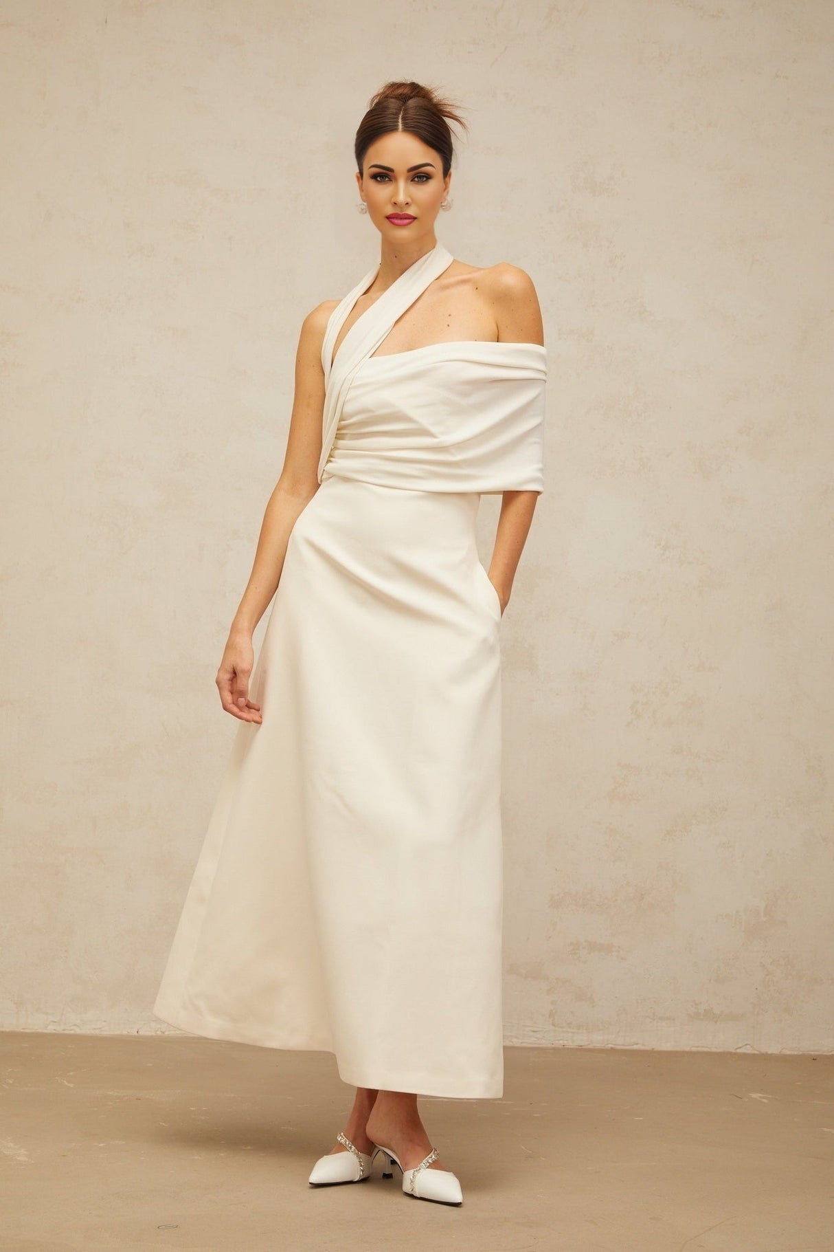A woman wearing a long creamwhite maxi dress with an asymmetric design standing in a neutral background