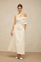 A woman wearing a long creamwhite maxi dress with an asymmetric design standing in a neutral background