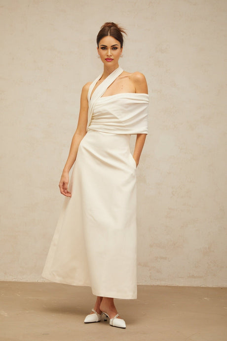 A woman wearing a long creamwhite maxi dress with an asymmetric design standing in a neutral background