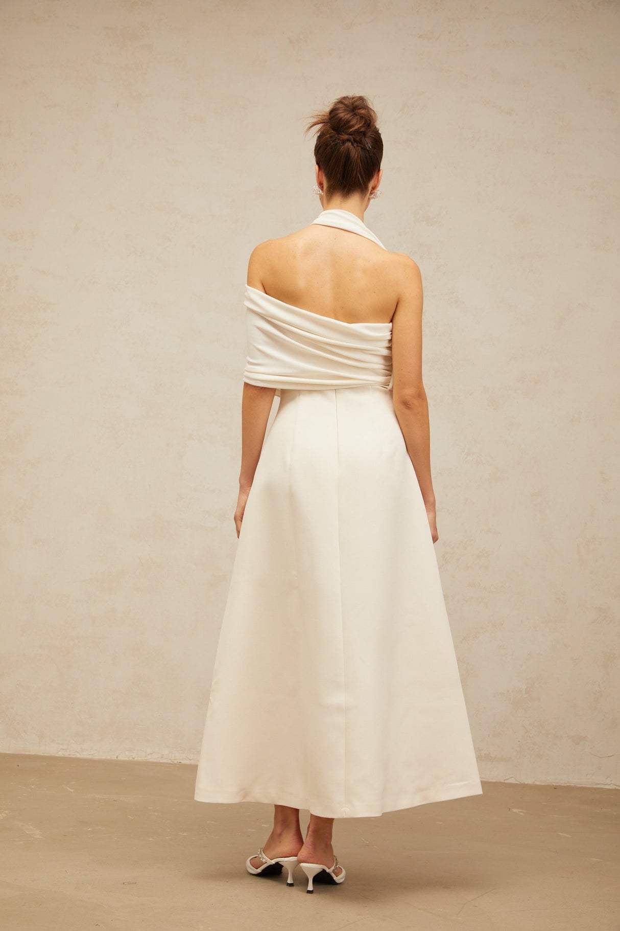 A woman wearing a long creamwhite maxi dress with an asymmetric hemline and neckline standing against a plain background