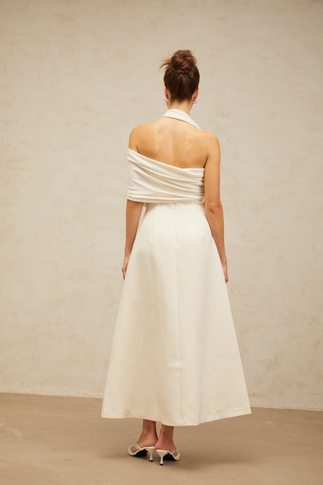A woman wearing a long creamwhite maxi dress with an asymmetric hemline and neckline standing against a plain background