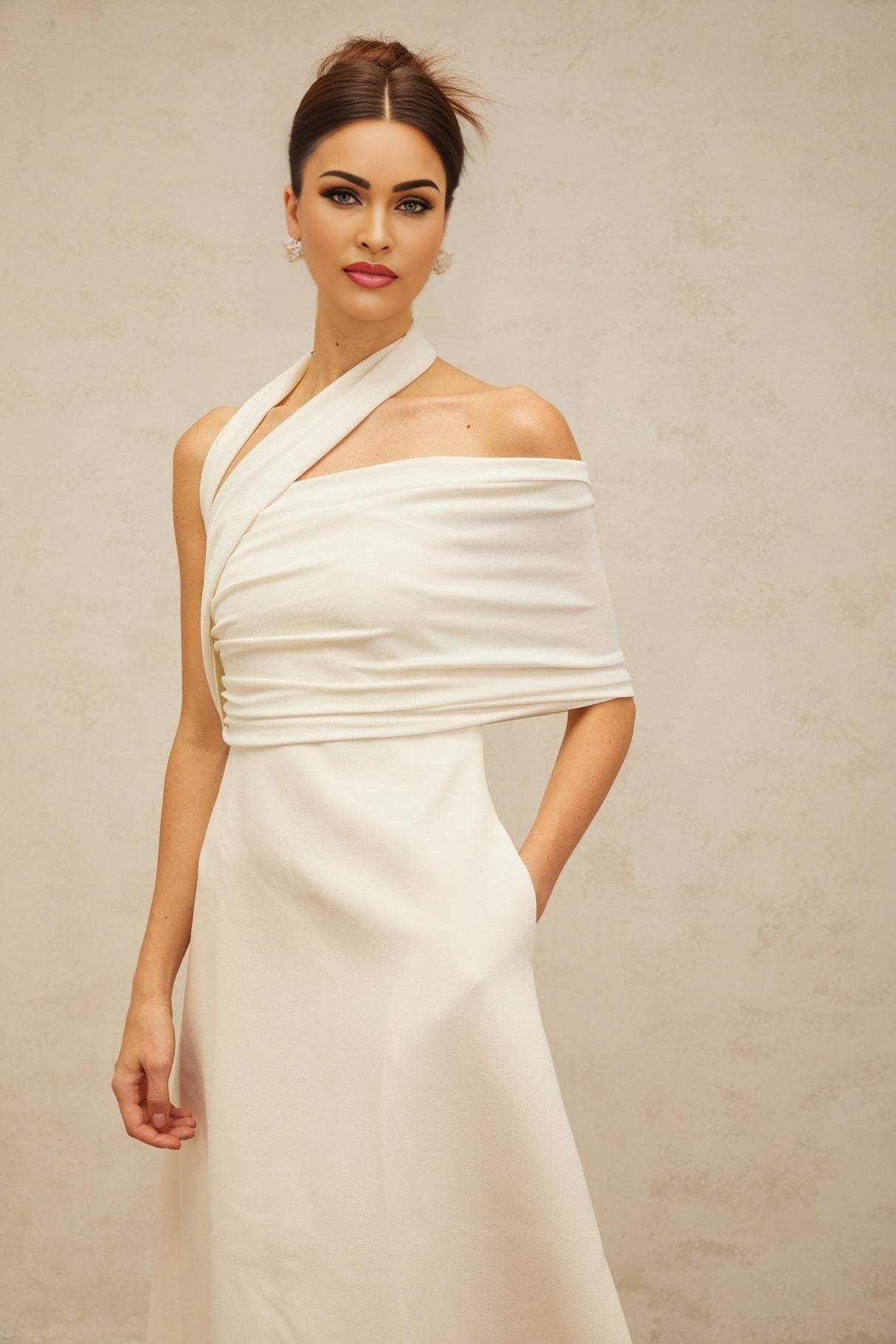 A woman wearing a long creamwhite maxi dress with an asymmetric neckline and hemline standing against a plain background