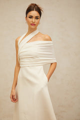 A woman wearing a long creamwhite maxi dress with an asymmetric neckline and hemline standing against a plain background