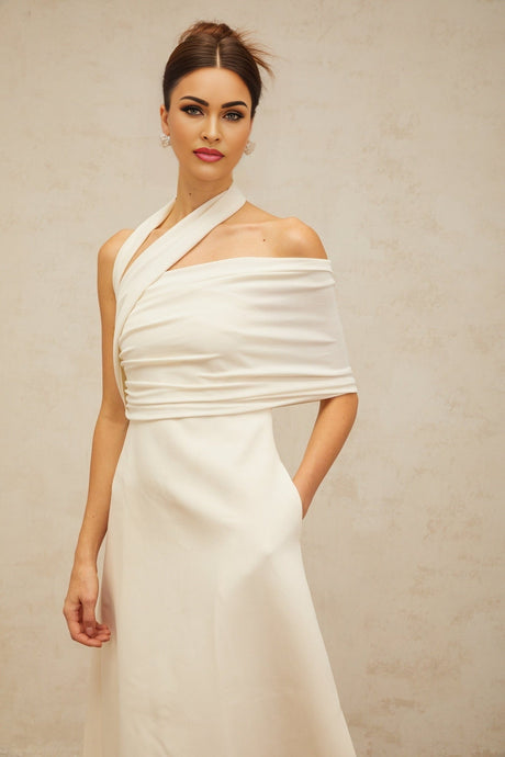 A woman wearing a long creamwhite maxi dress with an asymmetric neckline and hemline standing against a plain background