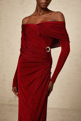 A woman wearing a long fitted red velvet dress with ruched detailing standing against a plain background