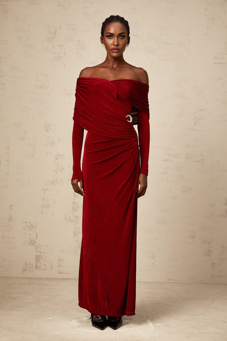 A woman wearing a long fitted red velvet dress with ruched detailing standing in an elegant pose