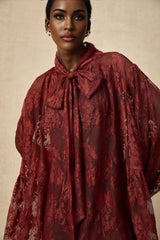 A woman wearing a long flowing red dress made of chantilly lace with an oversized fit standing against a plain background