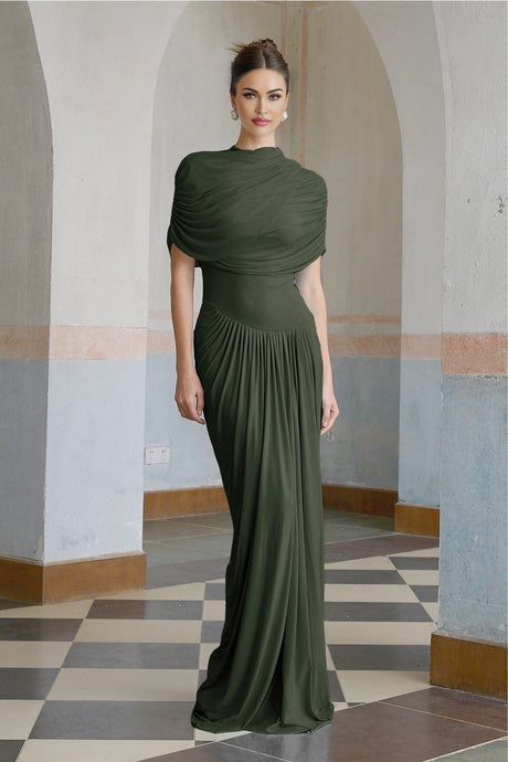 A woman wearing a long green asymmetric pleated maxi dress with a flowy silhouette and side slit standing against a plain background