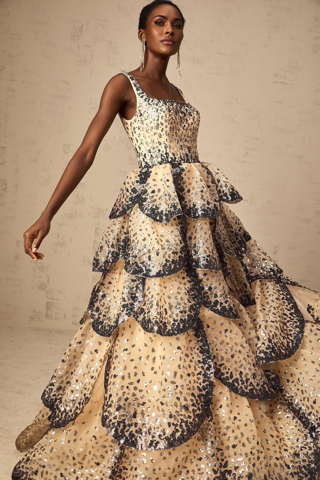 A woman wearing a long maxi dress with tiered layers and shimmering sequin details in a petal design standing in an elegant pose