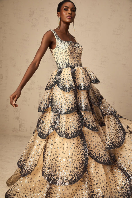 A woman wearing a long maxi dress with tiered layers and shimmering sequin details in a petal design standing in an elegant pose