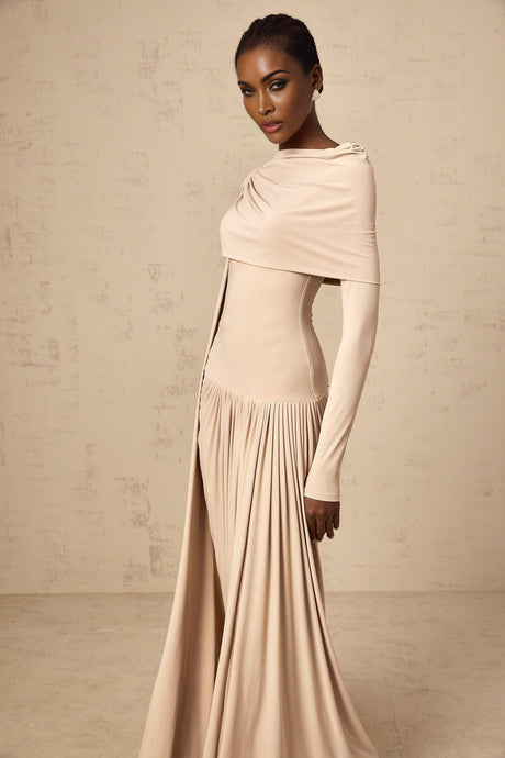 A woman wearing a long neutralcolored maxi dress with ribbon design details and elegant draping