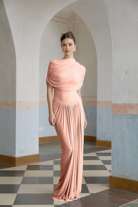 A woman wearing a long pink asymmetric pleated maxi dress with one shoulder strap standing against a plain background
