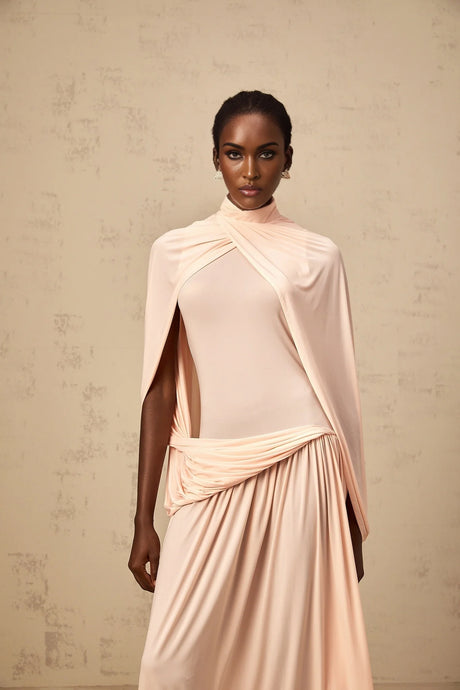 A woman wearing a long pink maxi dress with a flowing cape design and asymmetrical hemline