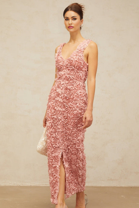 A woman wearing a long pink maxi dress with a Vneck and floral pattern standing in a relaxed pose against a plain background