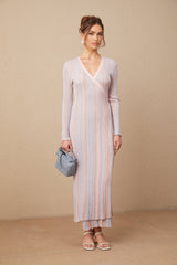 A woman wearing a long pleated knit dress with a V neckline in a soft color standing against a plain background