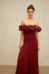 A woman wearing a long red knit maxi dress with a braided design detail standing against a plain background