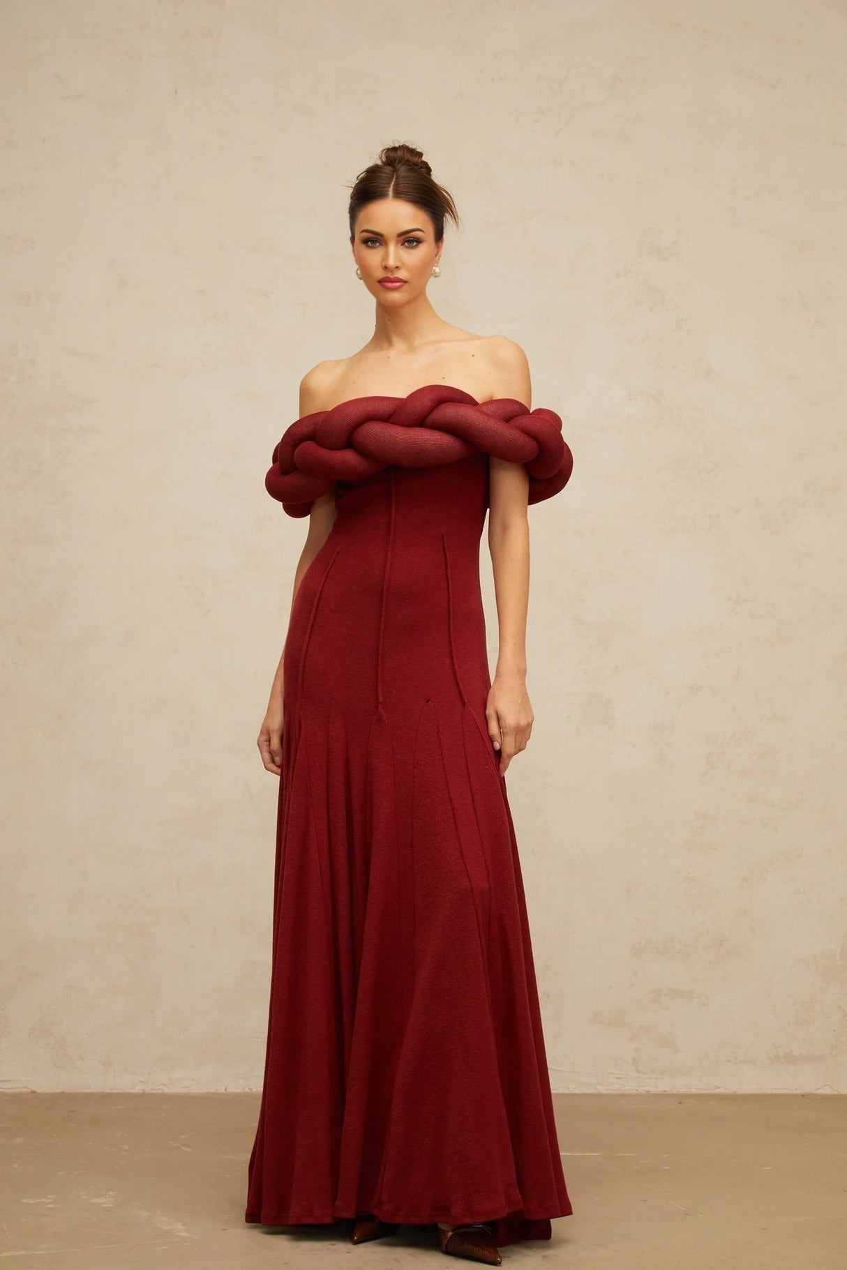 A woman wearing a long red knitted maxi dress with braided design details standing against a plain background