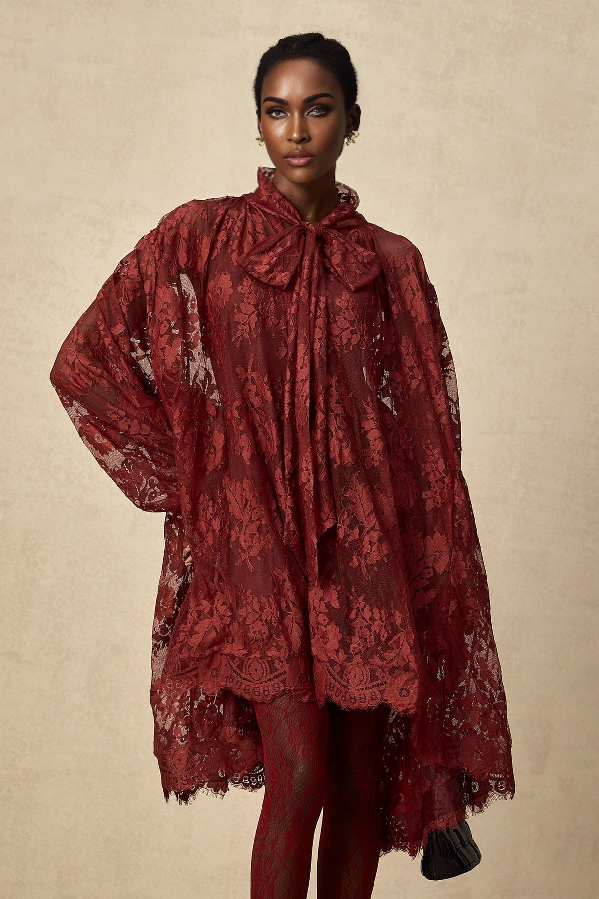 A woman wearing a long red lace dress with oversized sleeves and a flowy silhouette standing against a plain background