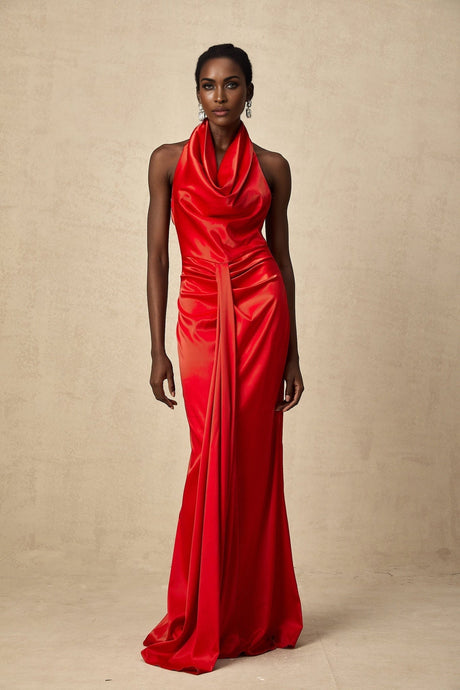 A woman wearing a long red satin dress with a halter neckline and draped details standing against a plain background