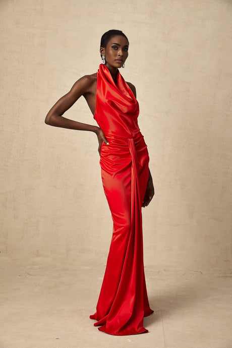 A woman wearing a long red satin dress with a halter neckline and draped fabric detail standing against a plain background