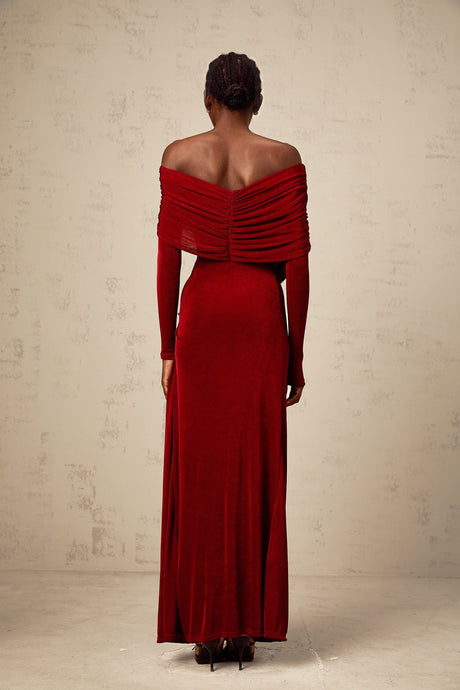 A woman wearing a long red velvet dress with ruched detailing and a fitted silhouette standing against a plain background