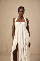 A woman wearing a long white chiffon maxi dress with a halter neck and ruffled details standing against a plain background