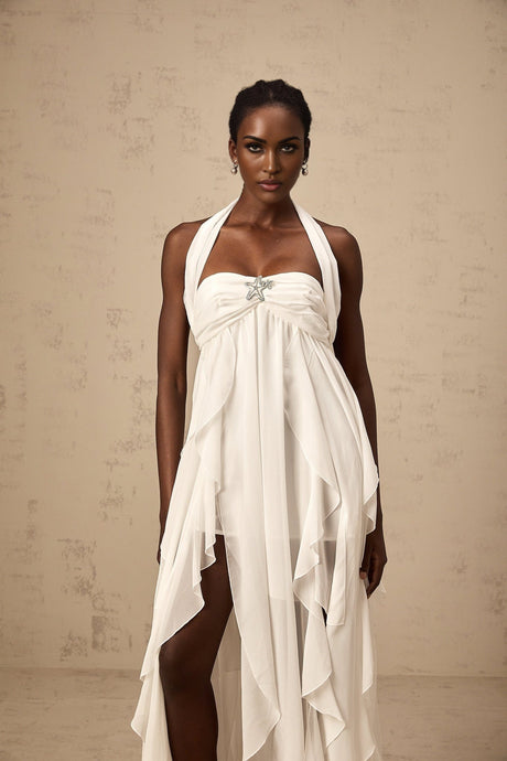 A woman wearing a long white chiffon maxi dress with a halter neck and ruffled details standing against a plain background