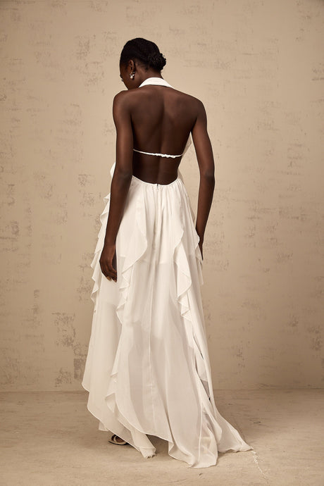 A woman wearing a long white chiffon maxi dress with ruffles and a halter neckline standing against a plain background