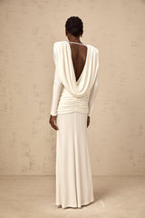 A woman wearing a long white fitted dress with ruched detailing standing against a plain background