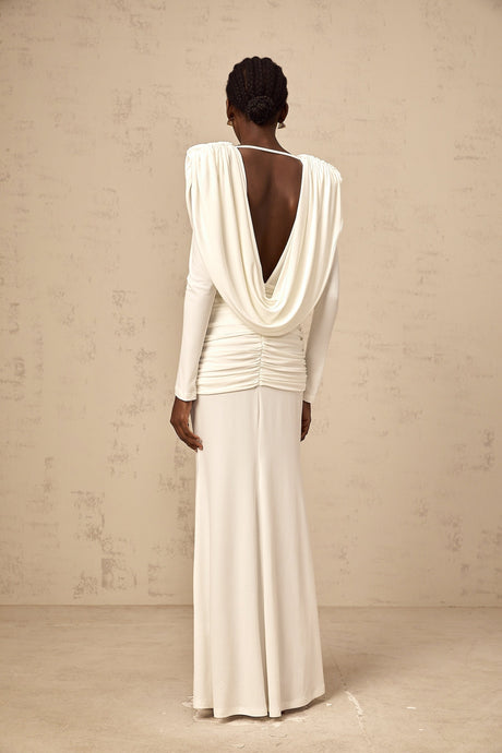 A woman wearing a long white fitted dress with ruched detailing standing against a plain background