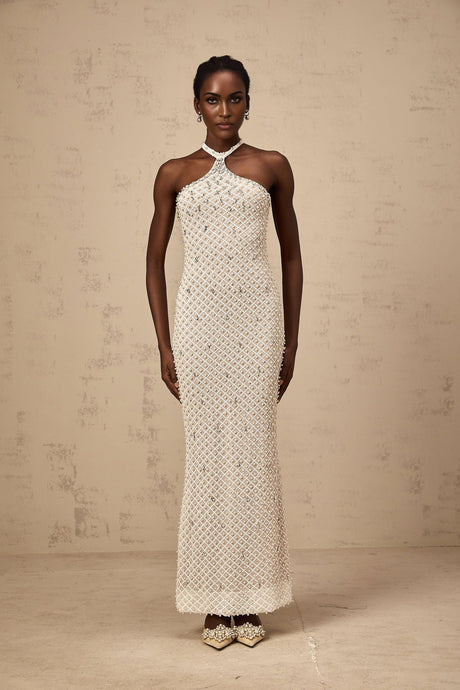 A woman wearing a long white halterneck maxi dress with embellishments standing against a plain background