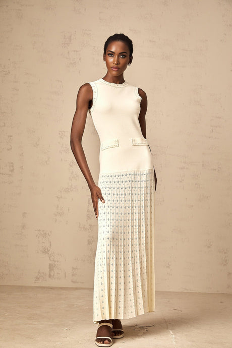 A woman wearing a long white knitted maxi dress with faux pearl embellishments standing in a neutral background