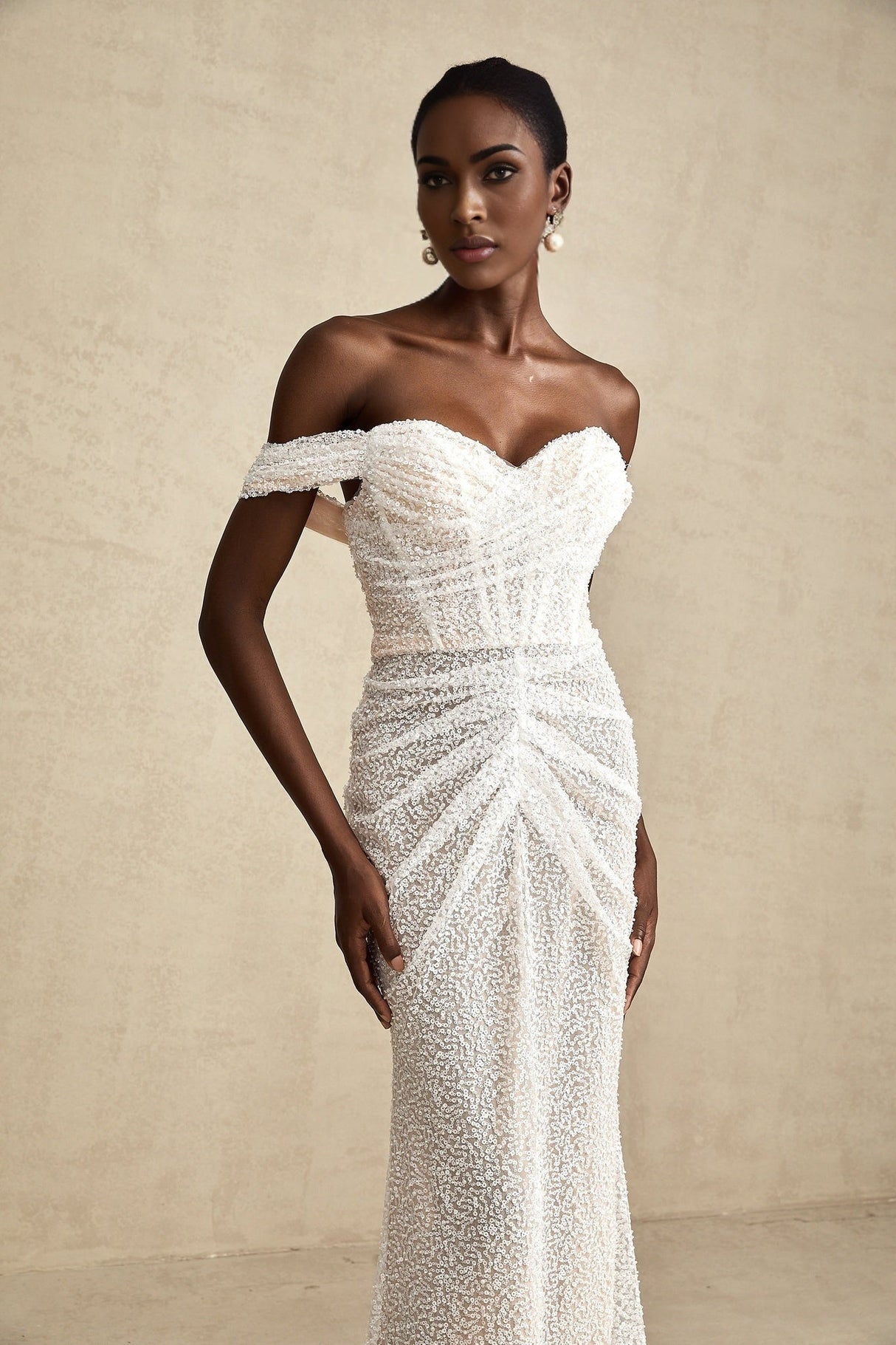 A woman wearing a long white offshoulder maxi dress with embellishments standing against a plain background