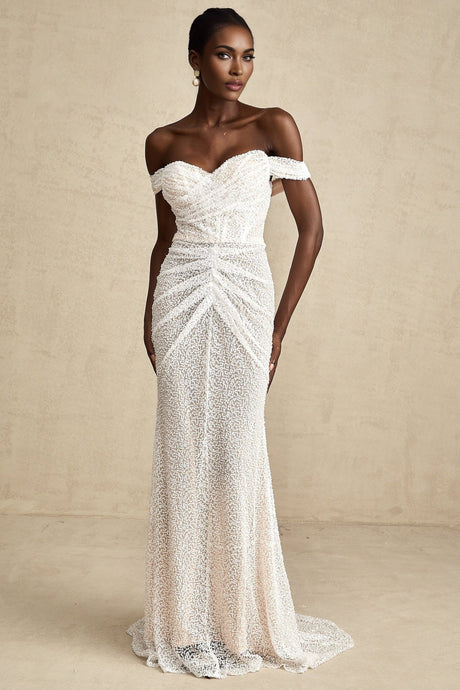 A woman wearing a long white offshoulder maxi dress with embellishments standing against a plain background