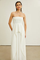 A woman wearing a long white offshoulder maxi dress with pleated details standing against a plain background