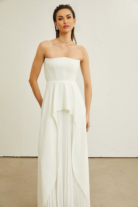 A woman wearing a long white offshoulder maxi dress with pleated details standing against a plain background