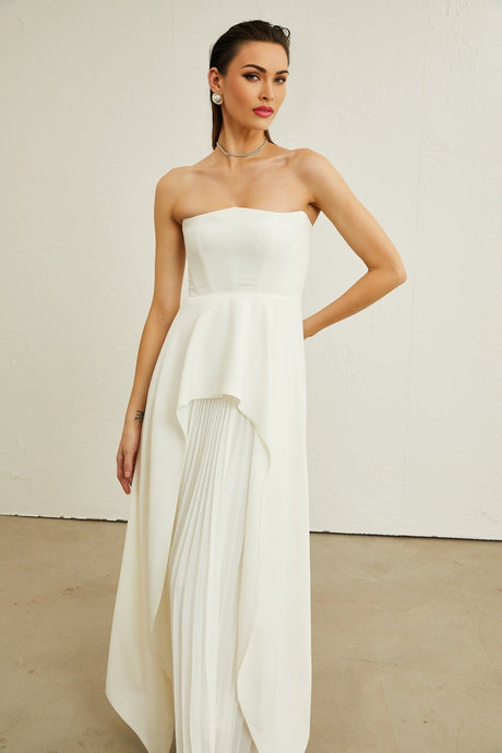 A woman wearing a long white offshoulder maxi dress with pleated details standing against a plain background