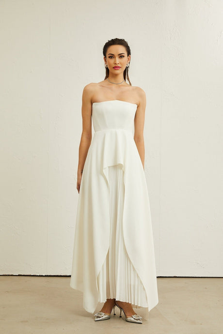 A woman wearing a long white pleated maxi dress with an offshoulder neckline posing against a plain background