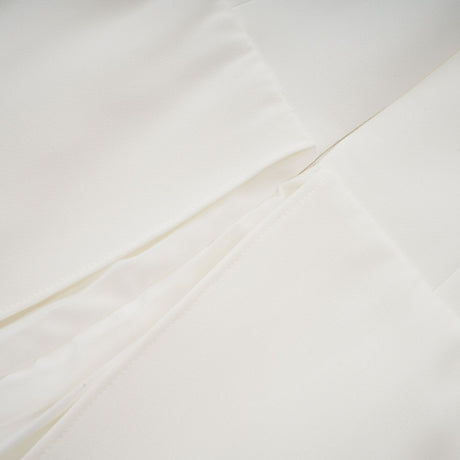 A woman wearing a long white pleated maxi dress with an offshoulder neckline standing against a plain background