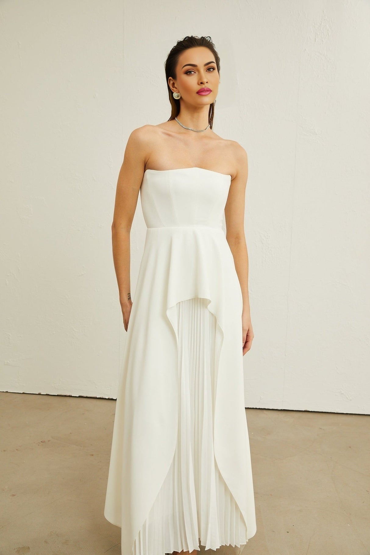 A woman wearing a long white pleated maxi dress with an offshoulder neckline standing against a plain background