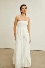 A woman wearing a long white pleated maxi dress with an offshoulder neckline standing against a plain background