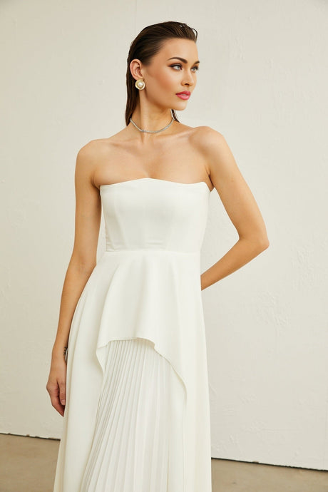 A woman wearing a long white pleated maxi dress with an offshoulder neckline standing against a plain background