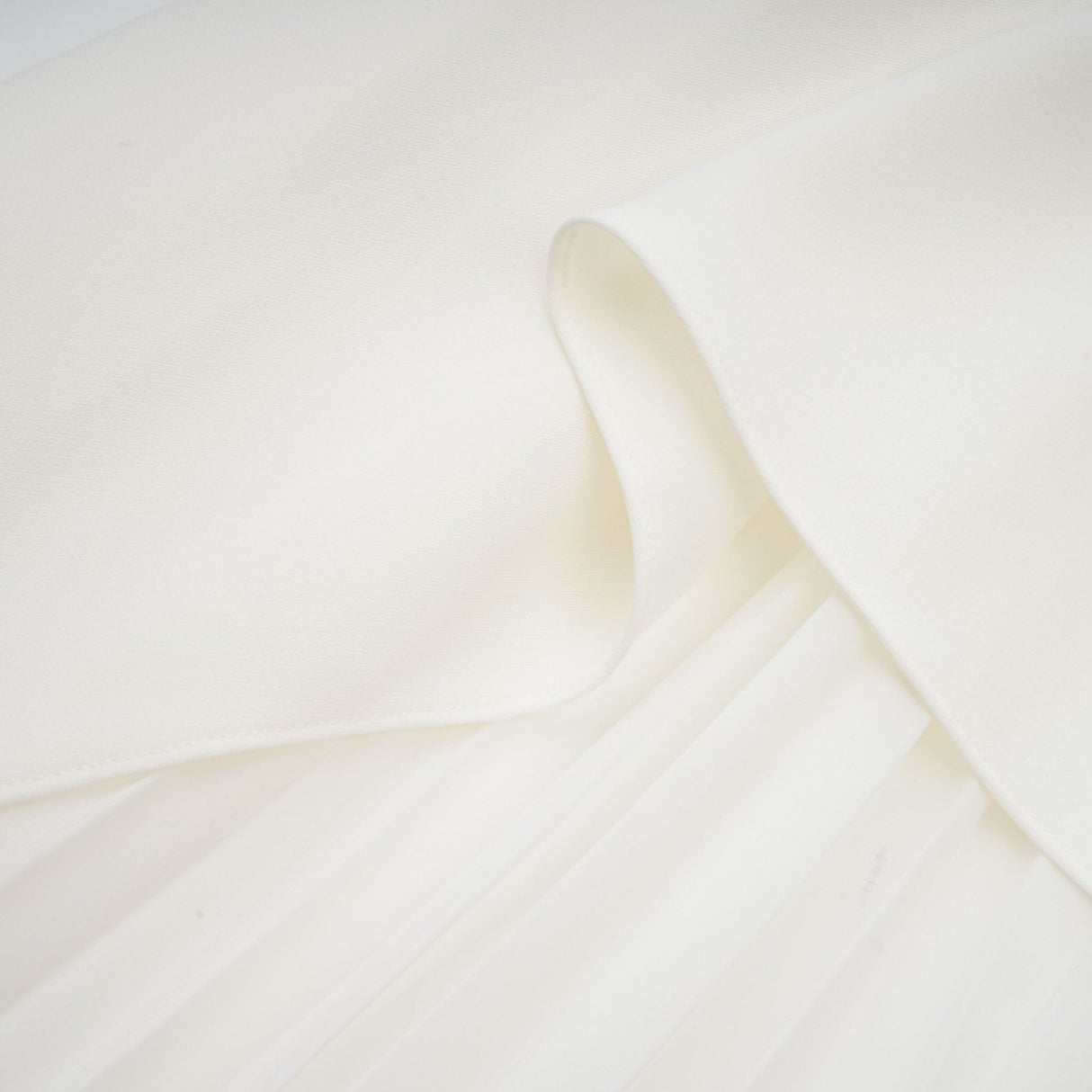 A woman wearing a long white pleated maxi dress with an offshoulder neckline standing in a relaxed pose