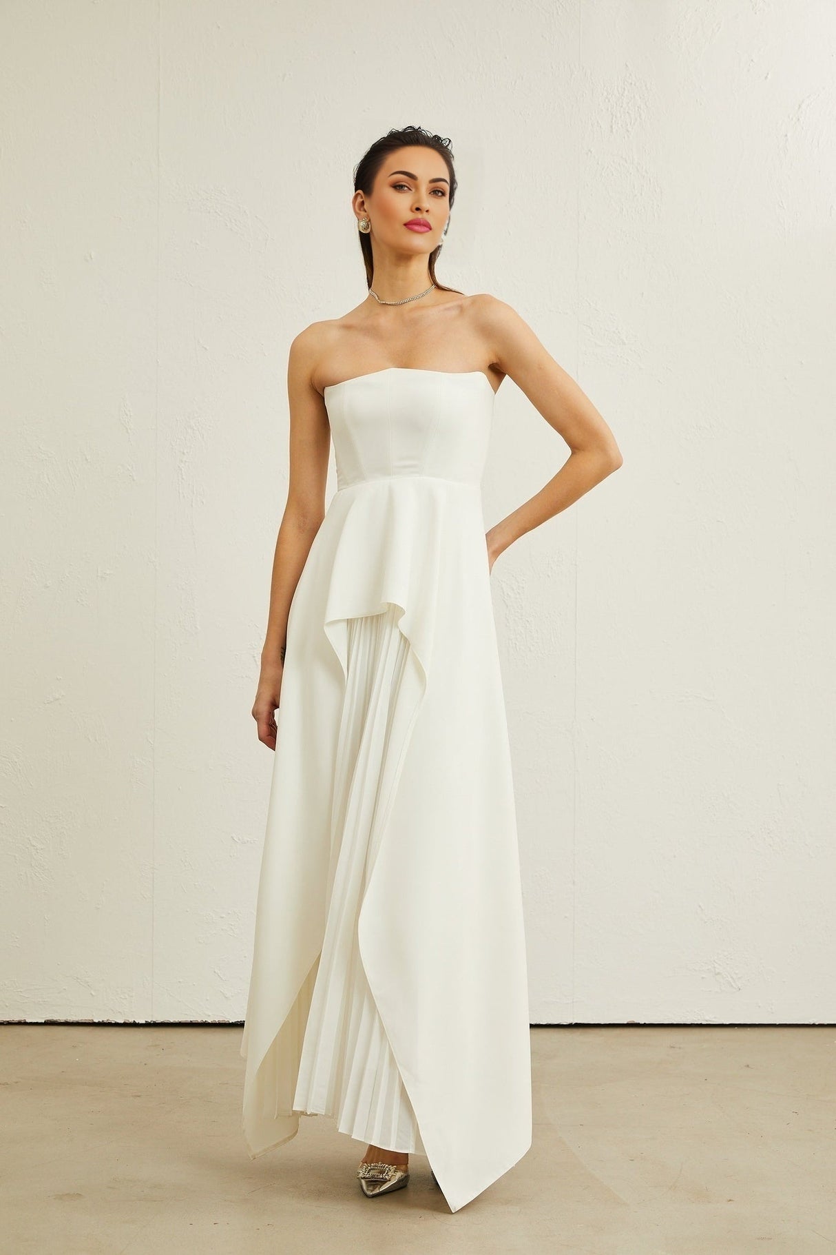 A woman wearing a long white pleated maxi dress with an offshoulder neckline