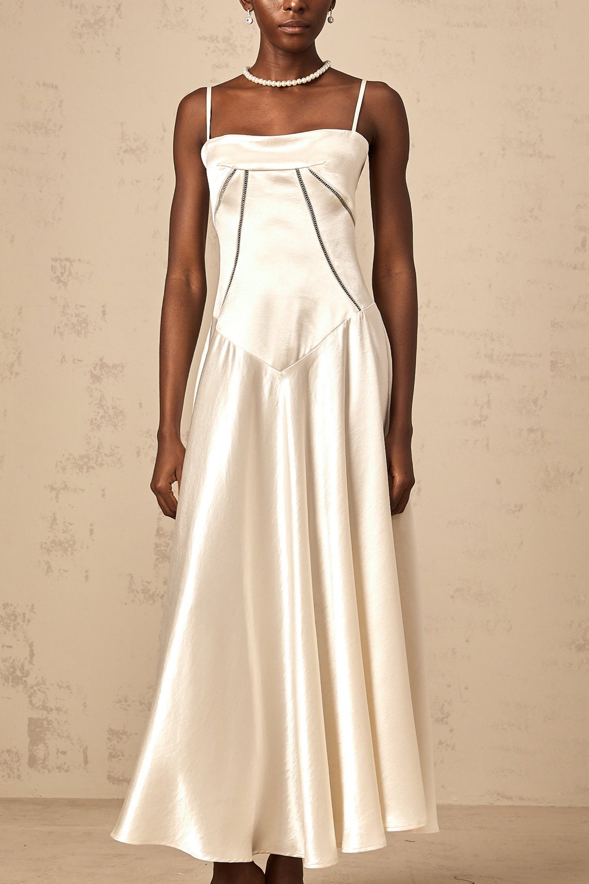 A woman wearing a long white satin dress with a flared skirt and fitted bodice standing against a plain background