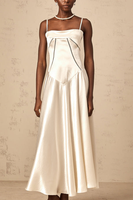 A woman wearing a long white satin dress with a flared skirt and fitted bodice standing against a plain background