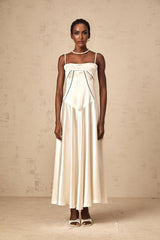 A woman wearing a long white satin dress with a flared skirt standing against a plain background