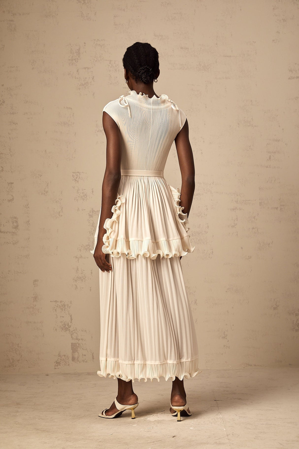 A woman wearing a long white sleeveless maxi dress with ruffle details and pleats standing against a plain background